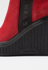 Czerwone zamszowe botki na koturnie z czarną elastyczną opaską z napisem „Calvin Klein Jeans”. Teksturowana czarna gumowa podeszwa z detalem marki.