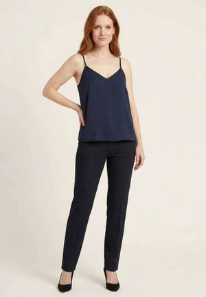 Mujer con cabello rojo que lleva puesta una camiseta sin mangas azul marino, pantalones negros ajustados y tacones negros, de pie con una mano en la cadera.