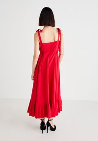 Woman with straight dark hair wearing a sleeveless red midi dress with tie straps and black high-heeled sandals, standing against a white background.
