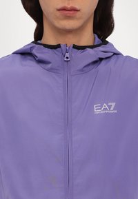 Casaco roxo com fecho éclair e capuz, de textura leve e suave; apresenta um acabamento preto e um logótipo EA7 do lado esquerdo.