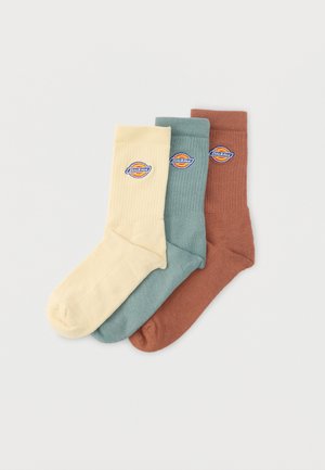 Trije zloženi Dickies nogavice za moške v bež, turkizni in rjavi barvi, vsaka z okroglim logotipom blagovne znamke na manšeti.