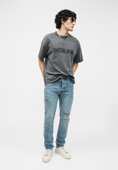 Νέος άνδρας που φοράει μαύρο φθαρμένο oversized t-shirt, ανοιχτό μπλε σκισμένο τζιν, λευκά αθλητικά παπούτσια, ασημένια αλυσίδα και γυαλιά ηλίου, στέκεται με τα χέρια πίσω από την πλάτη.