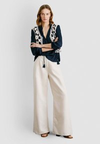 Mornarsko plava bluzica s čipkastim slojem i detaljima od resica, kombinirana sa širokim bež hlačama i smeškim sandalama. Glatka tekstura, opušten kroj.