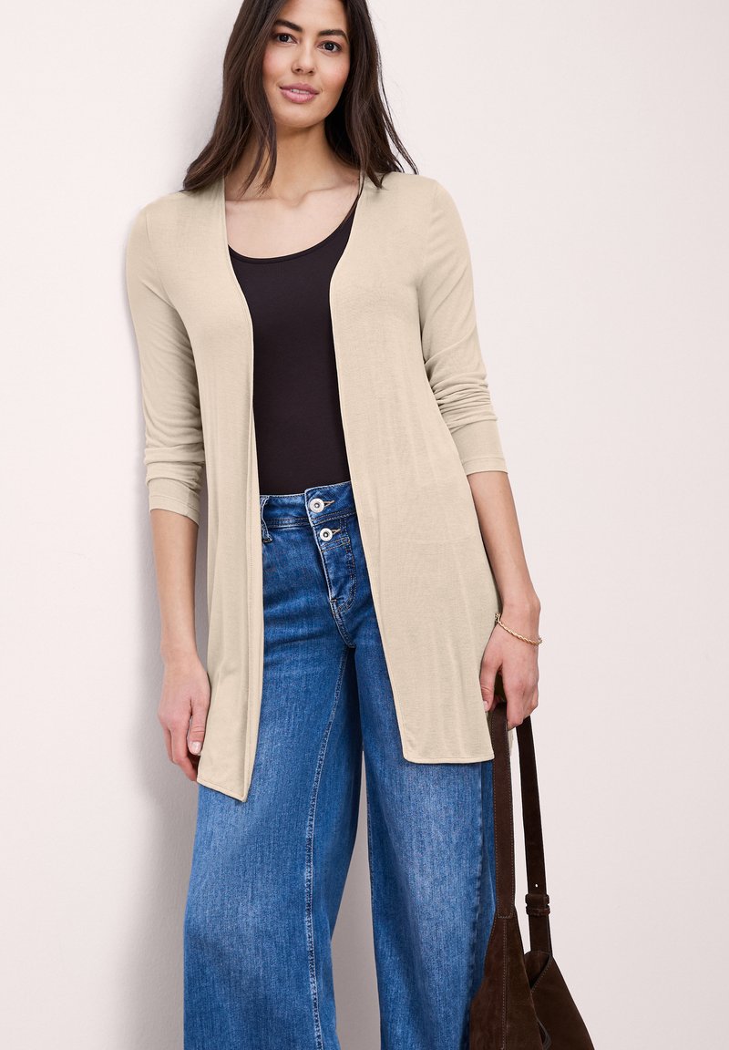 Femme portant un cardigan beige ouvert sur un haut noir et un jean large à taille haute bleu, tenant un sac à main marron foncé.