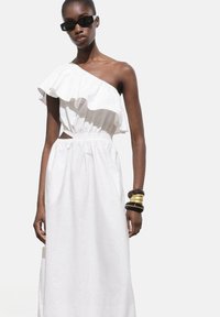 Robes maxi blanches à une épaule avec des volants et une taille élastique. Le mannequin porte des lunettes de soleil noires et des bracelets en or superposés.