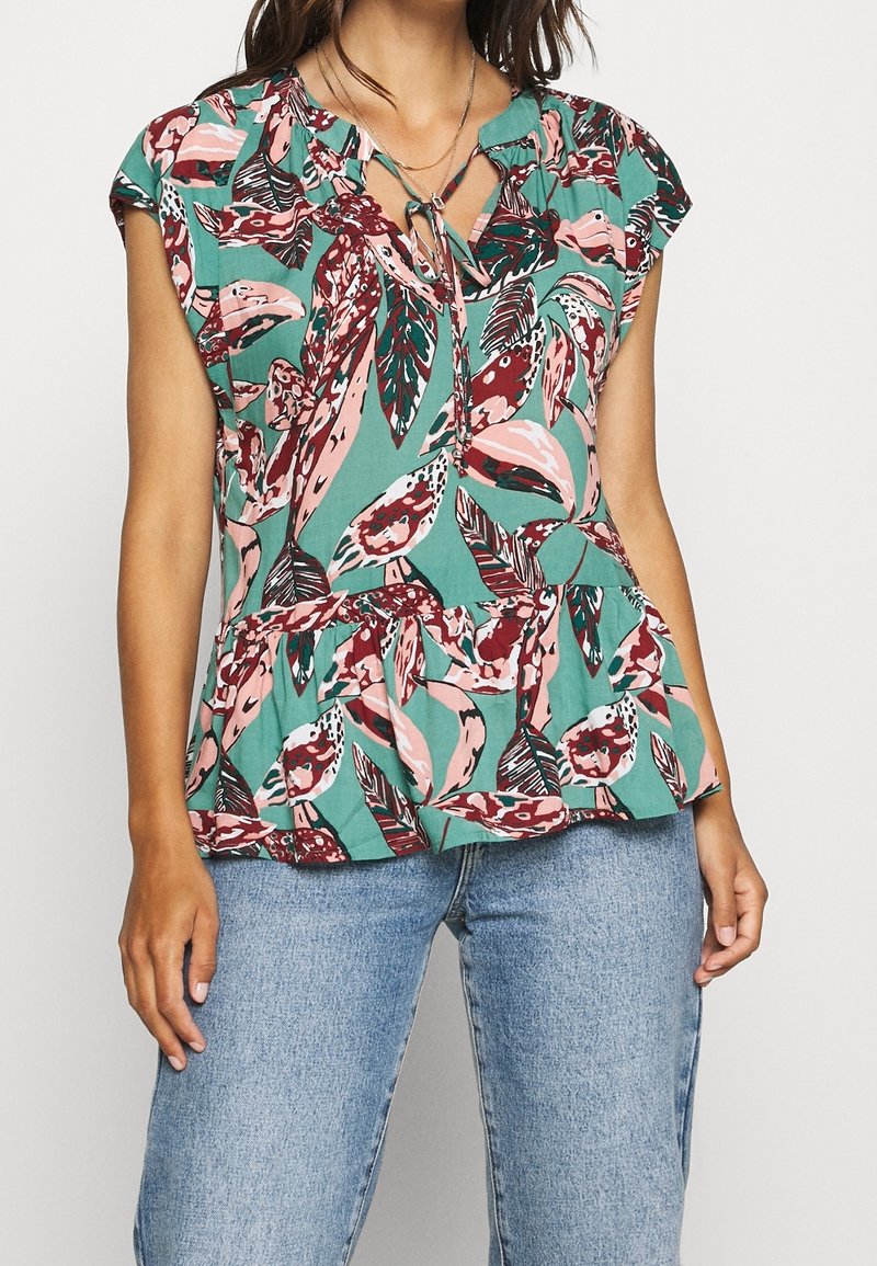 Kortærmet grøn bluse med et blomsterprint i pink og burgunder, som har en gathering peplum kant og en bindebånd ved halsen.