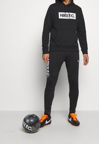 Sweat à capuche noir Nike avec logo blanc, assorti à un pantalon noir Nike F.C. avec logo blanc. Ballon de football noir avec accents orange.