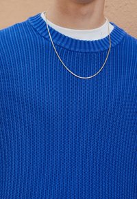 Un pull en maille bleu avec une texture côtelée et un col arrondi, superposé à une chemise blanche. Une fine chaîne en or repose sur le col.