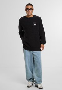 Maglione nero in maglia con texture a costine, collo rotondo e toppe sul petto. Abbinato a jeans larghi blu chiaro e sneakers bianche con accenti scuri.