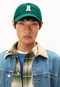 Grüne Kappe mit einem weißen Logo und gebogenem Schirm. Tragen mit einer Jeansjacke und einem karierten Hemd, was geschichtete Texturen und Farben zeigt.