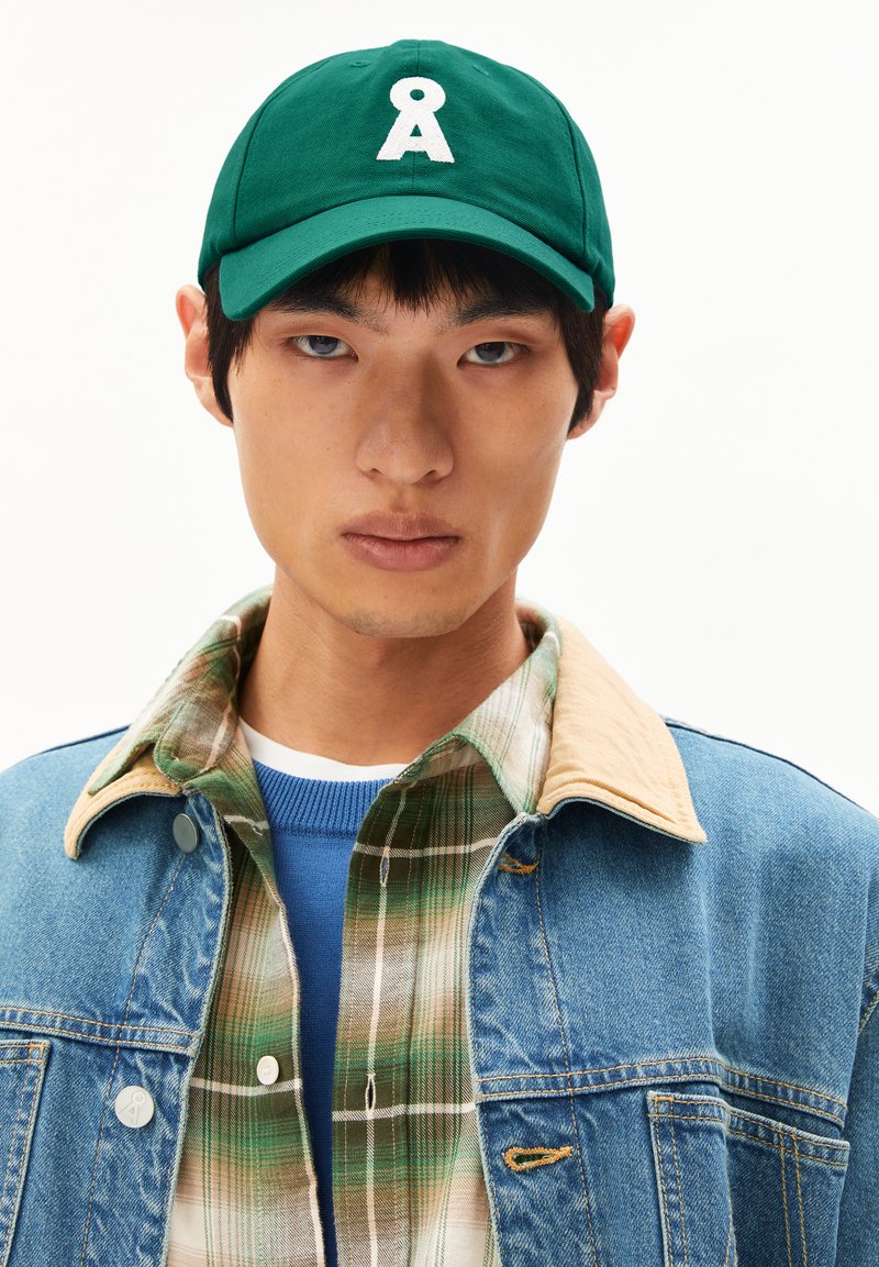 Grüne Kappe mit einem weißen Logo und gebogenem Schirm. Tragen mit einer Jeansjacke und einem karierten Hemd, was geschichtete Texturen und Farben zeigt.