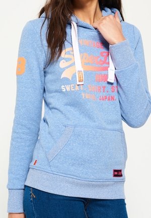 Woman in blue Superdry hoodie with orange and white logo, white drawstrings, and front pocket, wearing blue jeans.