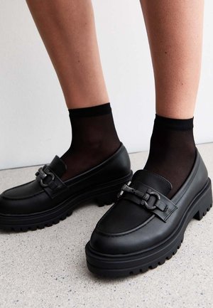 Jambes portant des socquettes noires transparentes et des mocassins noirs épais ornés d'une chaîne métallique décorative sur un sol clair.