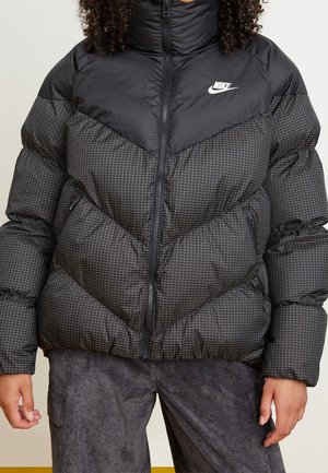 Vrouw draagt een zwart-grijs geruit Nike donsjack met ritszakken en grijze broek, staand met ontspannen handen.
