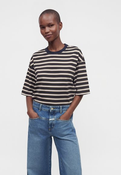 Personne debout, les mains dans les grandes poches d’un jean bleu, portant une chemise ample à manches courtes rayée noire et beige, faisant face à l’avant.