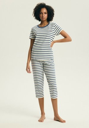 Kobieta stojąca boso, ubrana w biały i granatowo-biały paskowany krótki rękaw oraz spodnie do połowy łydki na jednolitym tle.