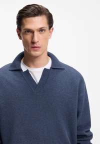 Blue wool sweater with a V-neck and ribbed collar design, layered over a white shirt. Smooth texture and relaxed fit.