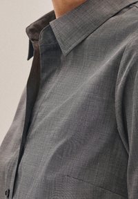 Grå mönstrad skjorta med knappar och en skarp krage, med fina linjer i ett strukturerat tyg. Innehåller en ficka på framsidan.