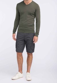 Olivgrüner V-Ausschnitt-Pullover, graue Cargo-Shorts mit Seitentaschen und weiße Sneaker. Das Outfit zeichnet sich durch ein figurnahes Design und weiche Texturen aus.