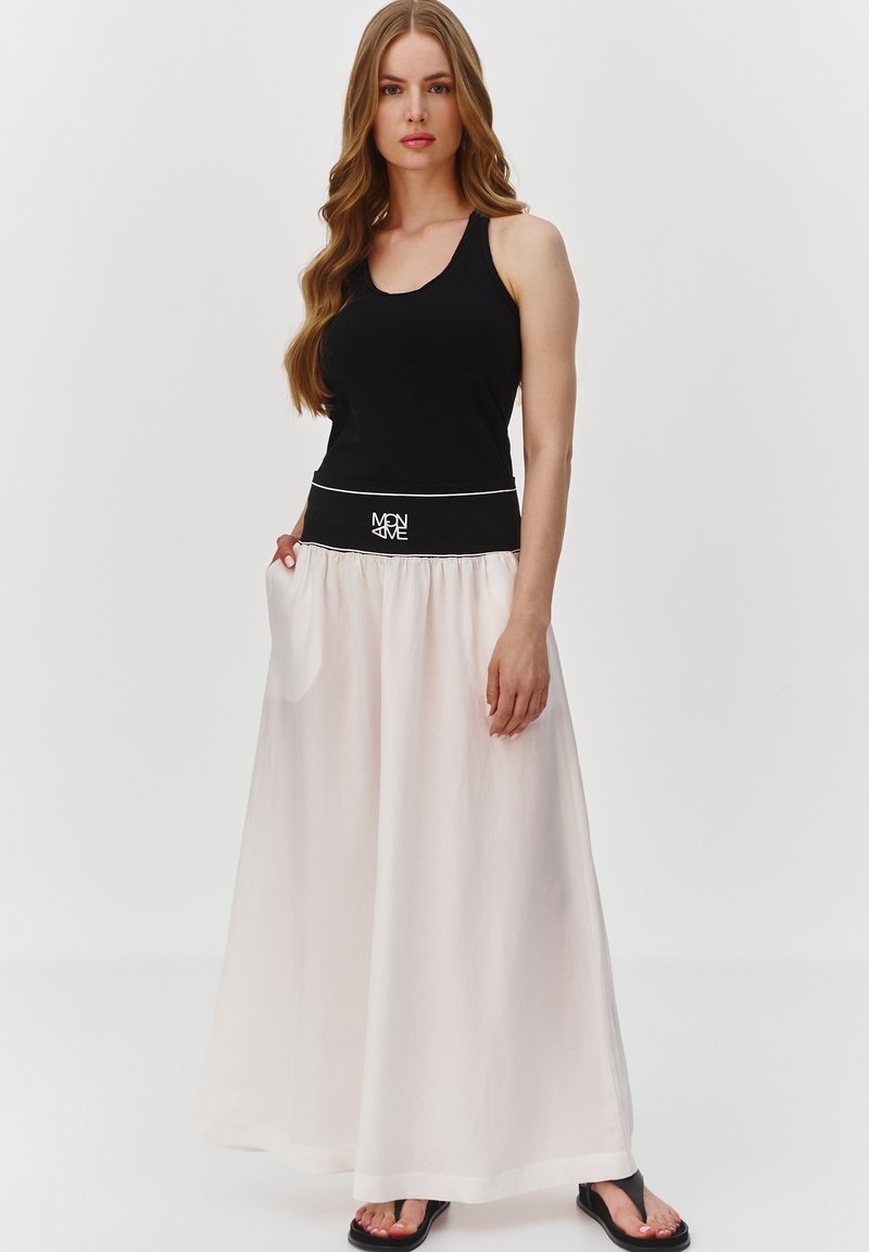 Czarny top na ramiączkach w połączeniu z luźną, białą spódnicą maxi, która ma czarny pas z białym wykończeniem i logo. Sandały dopełniają wygląd.