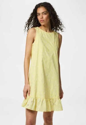 Femme aux cheveux bouclés portant une robe jaune sans manches avec des motifs floraux en broderie anglaise et un ourlet volanté, debout sur fond blanc.