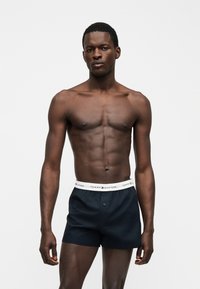 Μυώδης άνδρας με σκουρόχρωμο δέρμα, που φοράει σκούρο μπλε μποξεράκια Tommy Hilfiger με άσπρη μέση, στέκεται μπροστά σε απλό ανοιχτό φόντο.