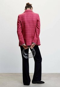 Vaaleanpunainen tweed-takki, jossa on kaulus ja fringeyksityiskohtia, yhdistettynä mustiin housuihin ja kirkkaaseen kaarevaan käsilaukkuun, nähtynä takaa.