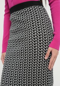 Frau trägt einen Rock mit schwarz-weißer geometrischer Musterung und schwarzem Bund sowie ein leuchtend pinkes Langarmoberteil, die Hand auf dem Rock ruhend.