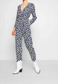 Marinblommig jumpsuit med långa ärmar, djup V-ringning, elastisk midja och sidofickor, kombinerad med vita klackade ankelskor.