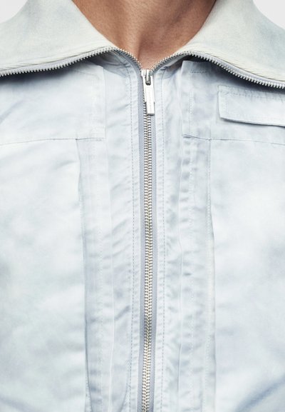 Κοντινό σε ένα άτομο που φοράει ανοιχτό γκρι μπουφάν με ένα κεντρικό ασημί φερμουάρ που είναι μερικώς φερμουάρ.