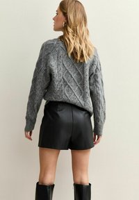 Grijze kabelgebreide trui met een ruime pasvorm, gecombineerd met zwarte leren shorts. De outfit bevat kniehoge zwarte laarzen en toont gestructureerde details.