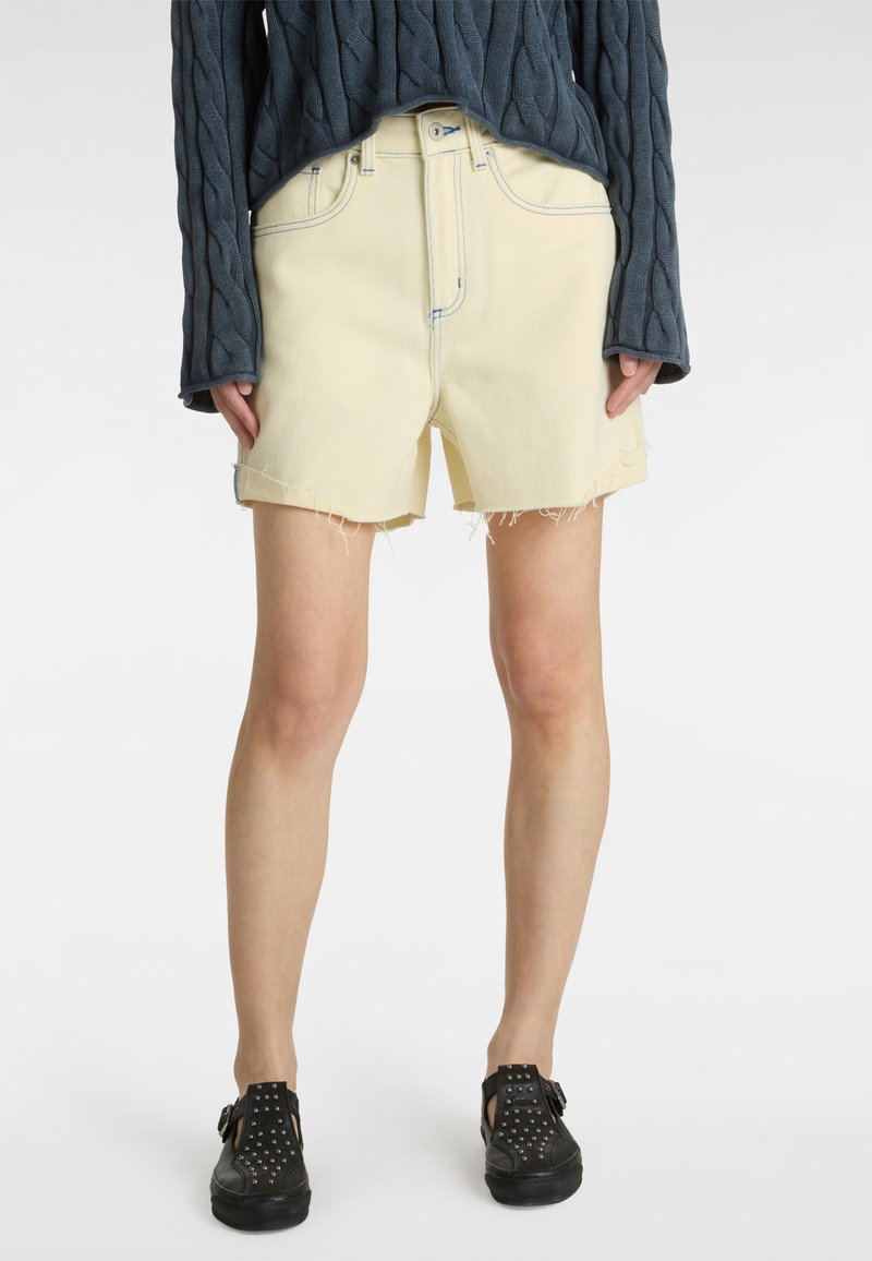 Shorts en denim jaune avec ourlets effilochés, taille haute et couture contrastée, associés à un pull en maille texturé bleu foncé et des chaussures noires à clous.