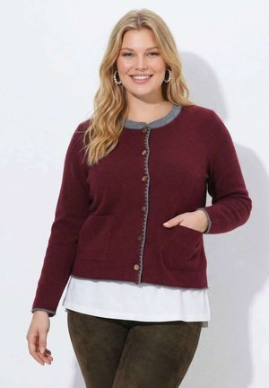 Donna sorridente con lunghi capelli biondi che indossa un cardigan bordeaux abbottonato sopra una maglietta bianca e pantaloni scuri, in posa con una mano in tasca.