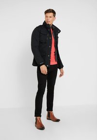 Black insulated jacket with button and zipper closure, multiple pockets. Paired with a coral shirt, black fitted pants, and brown chelsea boots.