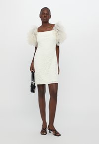 Witte kanten mini-jurk met een vierkante halslijn en doorschijnende beige tule pofmouwen. Het model draagt een kleine zwarte handtas en zwarte hakken.