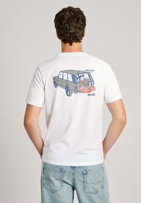 Camiseta blanca de algodón con un gráfico de un vehículo de color verde oliva con el maletero abierto, revelando artículos empaquetados. Diseño de corte relajado.