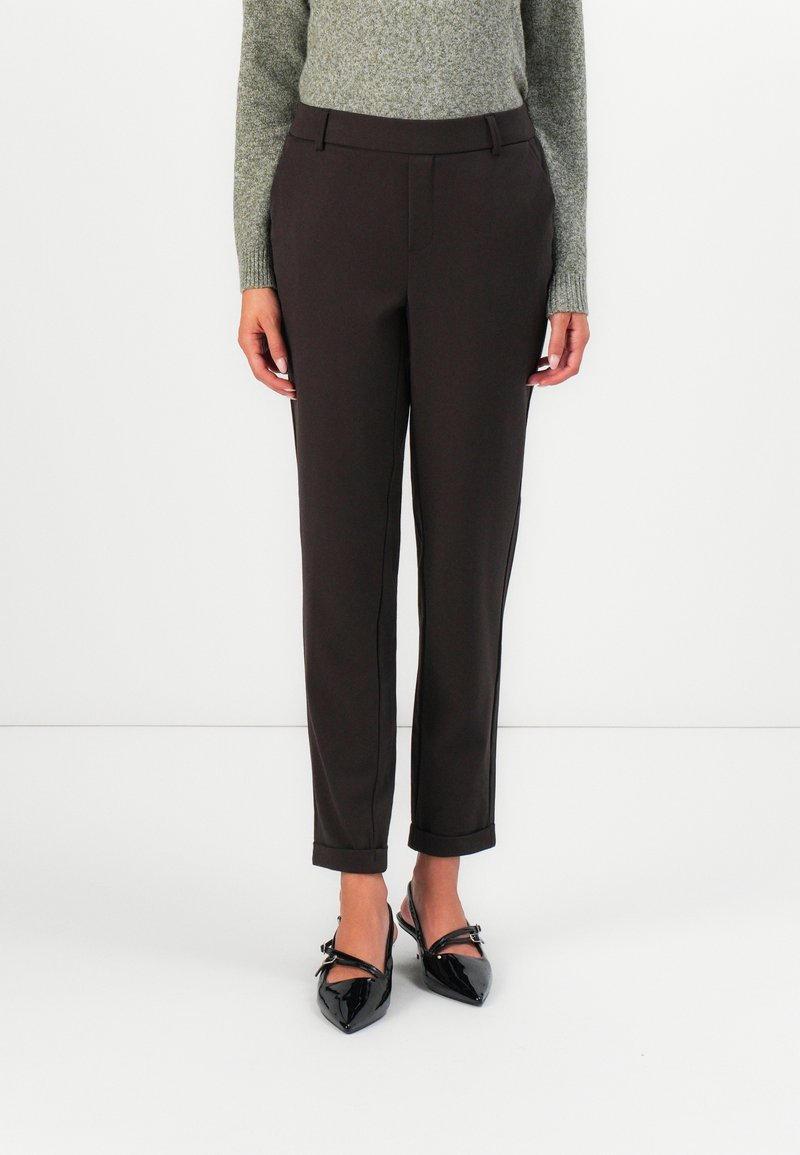 Femme portant un pantalon marron foncé coupé sur mesure, longueur cheville avec revers, un pull en maille gris et des escarpins noirs vernis à bout pointu avec des brides.