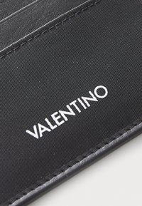 Portefeuille noir avec un tissu texturé et des accents en cuir. Le nom de la marque "VALENTINO" est affiché en lettres blanches.