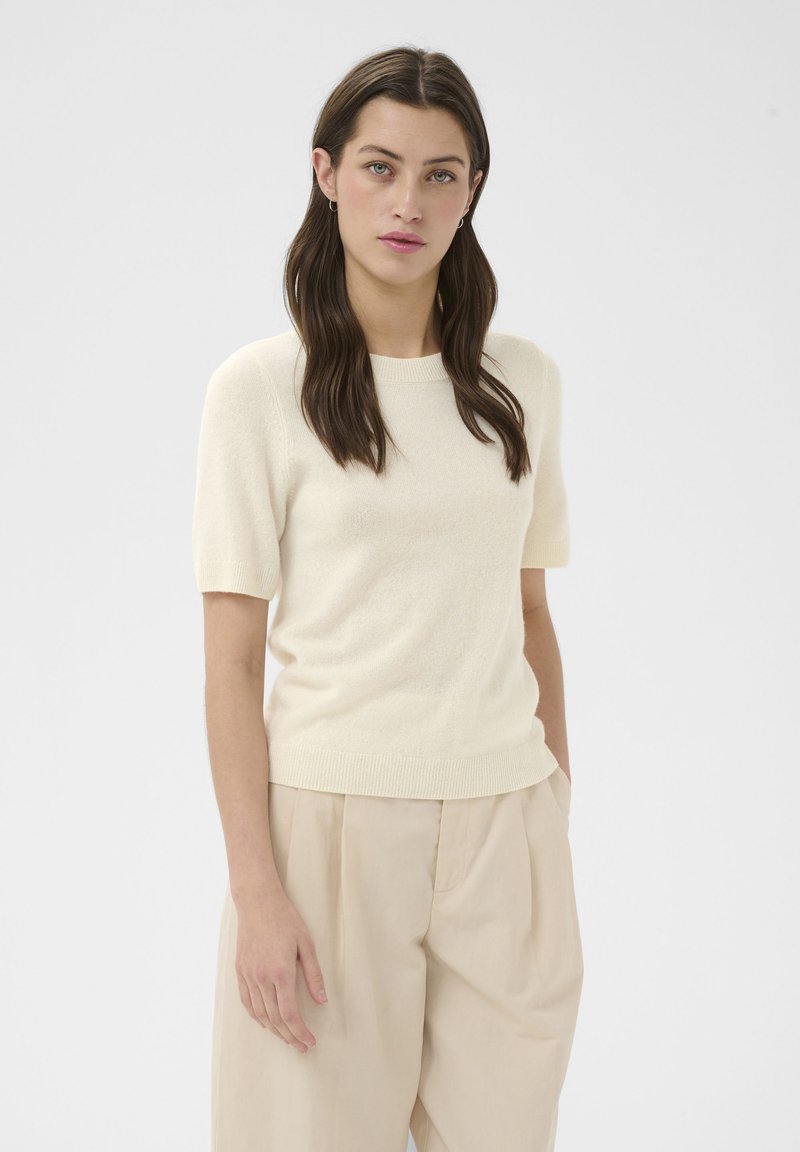 Femme aux longs cheveux foncés portant un pull crème à manches courtes et un pantalon beige à jambes larges, debout devant un fond uni.