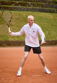 Bílá tenisová bunda Adidas na zip, černé šortky, hraje na antukovém hřišti, drží tenisovou raketu s růžovými akcenty. Viditelné krátké blond vlasy.
