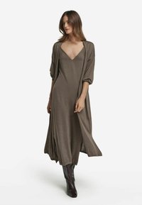 Lång, brun klänning med djup V-ringning, lösa ärmar och sidofickor, lager på lager med en matchande öppen cardigan. Bärs med mörka knähöga stövlar.