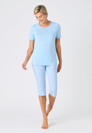 Frau geht barfuß, trägt ein hellblaues kurzärmeliges Oberteil und gemusterte Caprihosen vor einfachem Hintergrund.