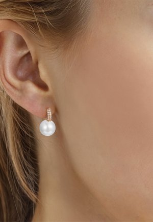 Pendiente de aro de oro con pequeños diamantes incrustados que sostienen una gran perla blanca en el lóbulo de la oreja de una mujer con cabello rubio.