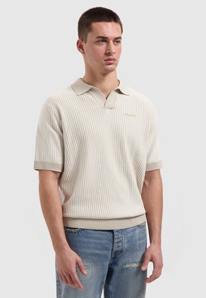 Jonge man die een beige, kort mouw, gestructureerd gebreid poloshirt draagt en lichtblauwe gescheurde jeans, staand tegen een effen achtergrond.