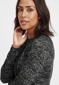 T-shirt long à rayures noir et blanc avec un col rond et un tissu texturé. Le modèle tient sa main près de son visage.