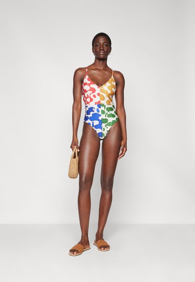 Costum de baie integrale colorat, cu un model de forme abstracte în nuanțe de roșu, portocaliu, albastru și verde. Modelul ține o geantă țesută și poartă sandale bej.