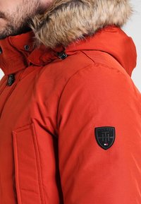 Muž v jasně oranžové zimní bundě s kapucí lemovanou kožešinou a černou nášivkou s logem Hilfiger na rukávu, detailní boční pohled.