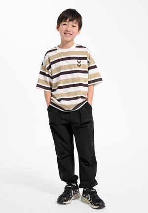 Smilende dreng iført beige og brune stribede T-shirt, sorte bukser og sorte sneakers, står med hænderne i lommerne på hvid baggrund.