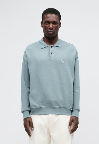 Ljusblå-grön långärmad pikétröja med krage, som har tre knappar och en liten logotyp på vänster bröst. Slät textur.