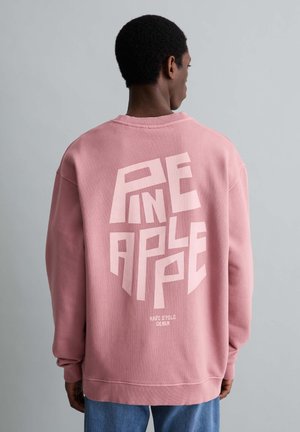 Homme portant un sweat-shirt rose avec un grand texte "PINEAPPLE" en rose clair et un texte plus petit "MARC O'POLO DENIM" au dos, tourné vers l'arrière.